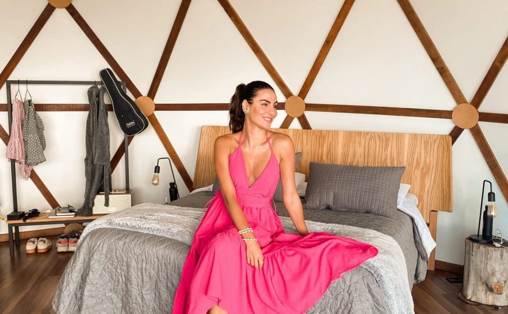 Mel Fronckowiak de vestido cor de rosa sentada em cama de quarto de hotel
