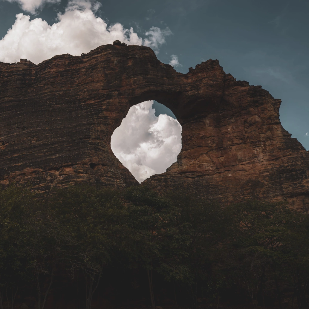 Fotografia em close na pedra furada da Serra da Capivara. A formação rochosa possui coloração escurecida com resquícios de alaranjado e tem uma abertura redonda no meio, por onde é possível ver o céu com nuvens.