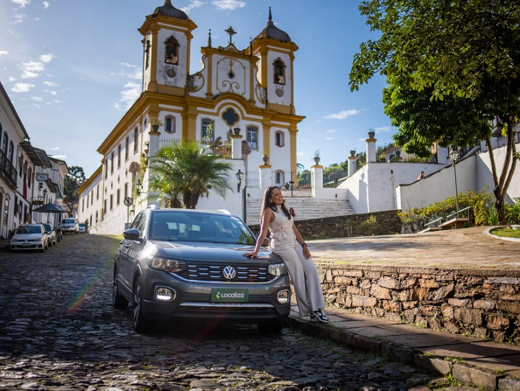Mulher encostada em um carro em uma ladeira de paralelepípedo da cidade histórica de Ouro Preto, em Minas Gerais. De fundo, uma das igrejas antigas da cidade.