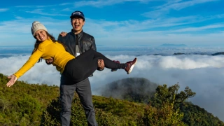 Casal posa para foto com homem segurando mulher no colo, no topo de uma montanha com vegetação e nuvens ao fundo