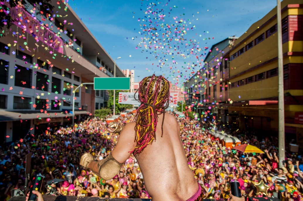 Bloco de rua em Belo Horizonte. Destaque para um artista em cima do trio, de costas para a câmera. Confetes coloridos caindo sobre a multidão.