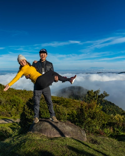 Casal posa para foto com homem segurando mulher no colo, no topo de uma montanha com vegetação e nuvens ao fundo