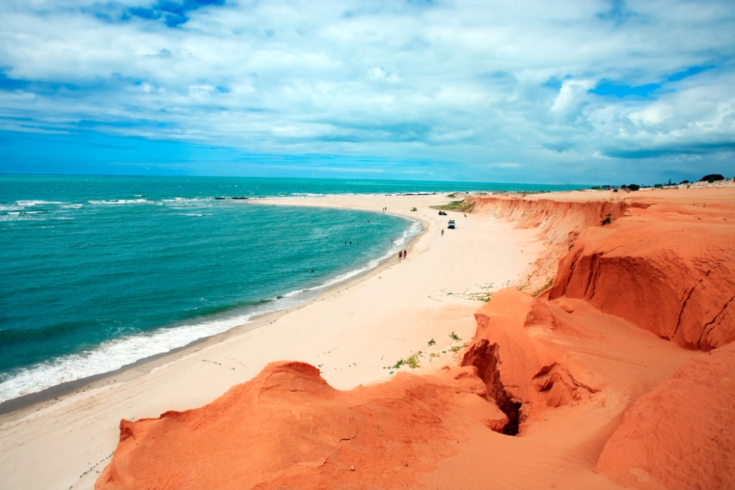Vista aérea de praia com mar verde-esmeralda, areia clara e falésias de cor laranja vibrante