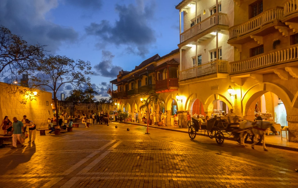 Praça no centro de Cartagena, ao anoitecer. O local está ocupado por pessoas sentadas nos bancos, e as luzes dos estabelecimentos estão acesas. No canto direito, há uma charrete puxada por um cavalo.