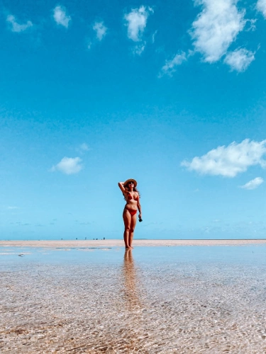 Uma mulher de trajes de banho caminha por bancos de areia em frente à uma praia deserta em dia ensolarado