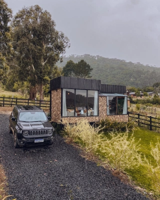 Paisagem de carro preto ao lado de pequena casa em região arborizada em dia nublado