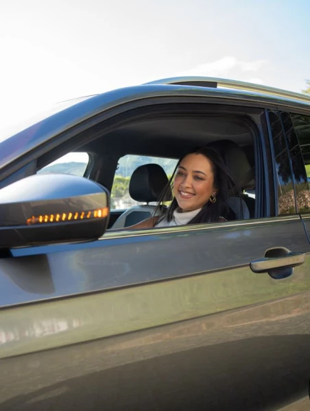 Mulher sorridente dirigindo um carro cinza escuro e olhando para o retrovisor que tem uma luz acesa num dia claro ilustrando as vantagens de locação de automóvel e quanto custa alugar um carro.