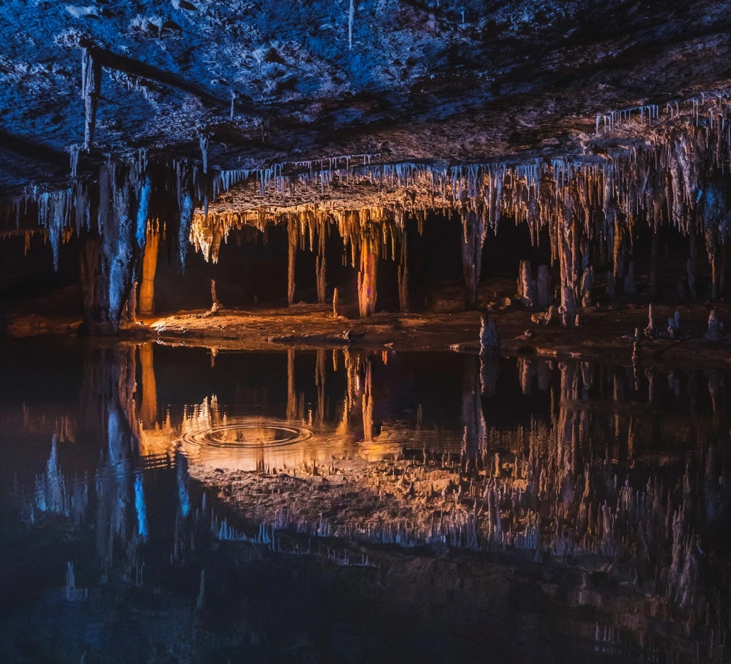 Interior de uma caverna com formações rochosas pontiagudas que refletem na água represada com cores douradas e azuis devido à iluminação externa