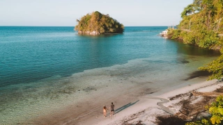 Vista aérea de uma praia e um casal de mãos dadas sobre a areia, avistando uma pequena ilha em meio ao mar