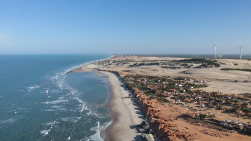 Vista aérea de praia com extensa faixa de areia, falésias avermelhadas e dunas de areia ao fundo