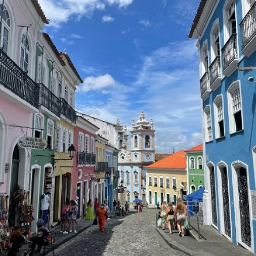 Pessoas caminham pelas ruas da cidade de Salvador com várias casas e construções coloridas e em estilo colonial com um dia ensolarado