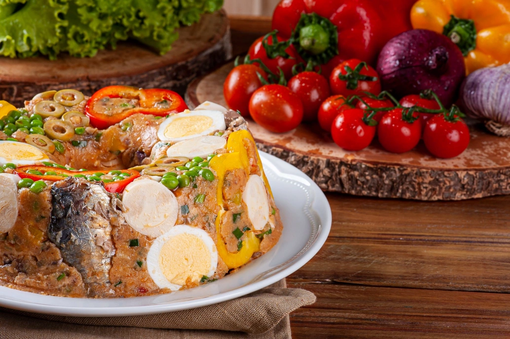 Em primeiro plano, um prato branco com cuscuz paulista, decorado com ovos, pimentões, ervilhas, sardinhas e azeitonas. Ao fundo, uma tábua com legumes frescos como tomates-cereja, cebolas e pimentões.