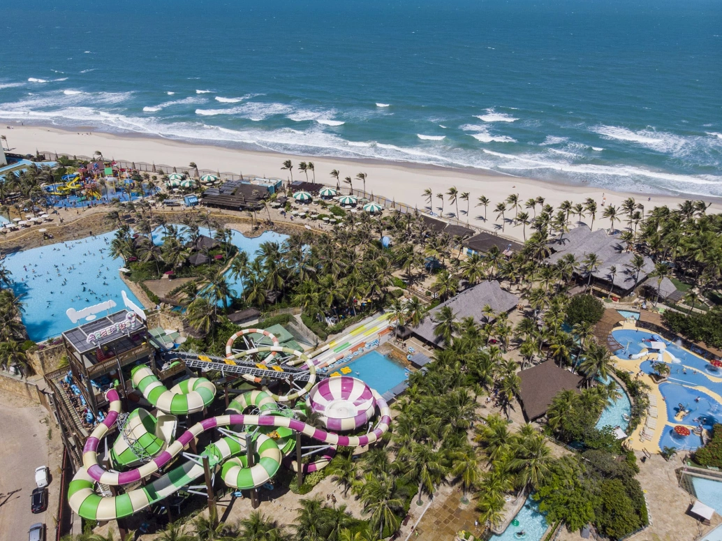 Imagem aérea de um parque aquático com tobogãs coloridos, com destaque à vegetação em volta e uma praia ao fundo.