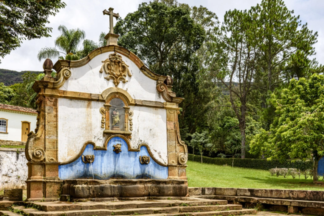 Fonte de água do século XVIII, em estilo barroco, em meio a uma praça arborizada em cidade histórica de Minas Gerais.