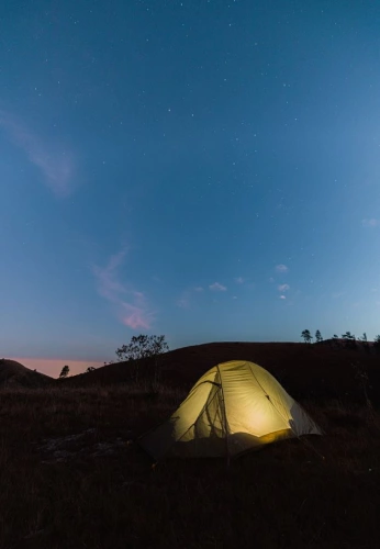 Barraca de acampamento com luz acesa dentro ao anoitecer, mostrando o céu parcialmente estrelado
