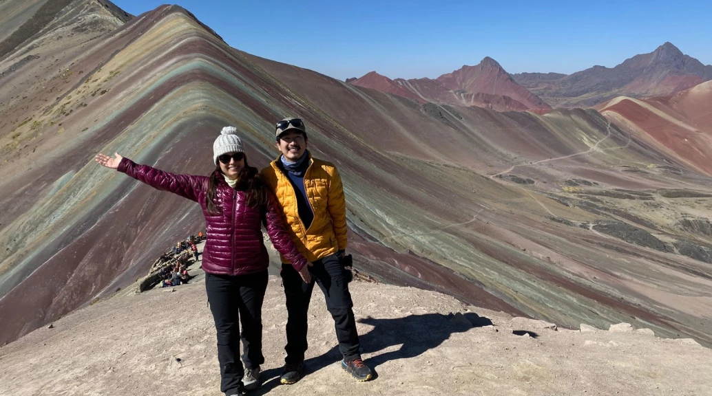 Mulher e homem em pé com grande uma montanha colorida ao fundo, na região de Ausangate, no Peru.