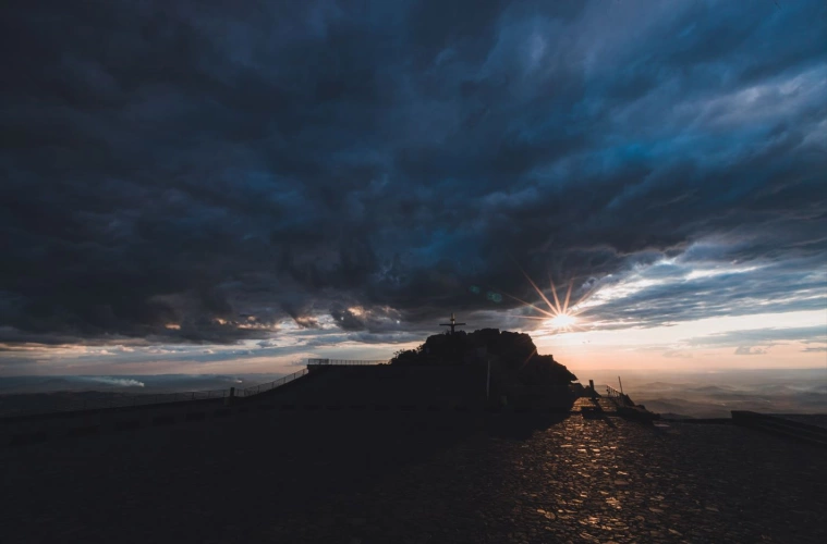 Feixes de luz solar entre nuvens marcando a silhueta de uma pedra e uma cruz no alto de uma colina