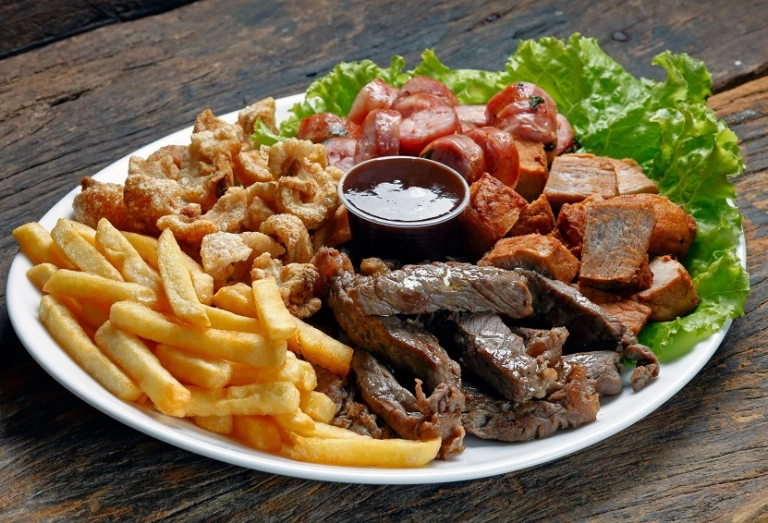 Um prato branco em uma mesa de madeira escura com batatas fritas, carne de boi, torresmo, carne de porco, linguiça e decorado com alface. Ao meio, um molho escuro.