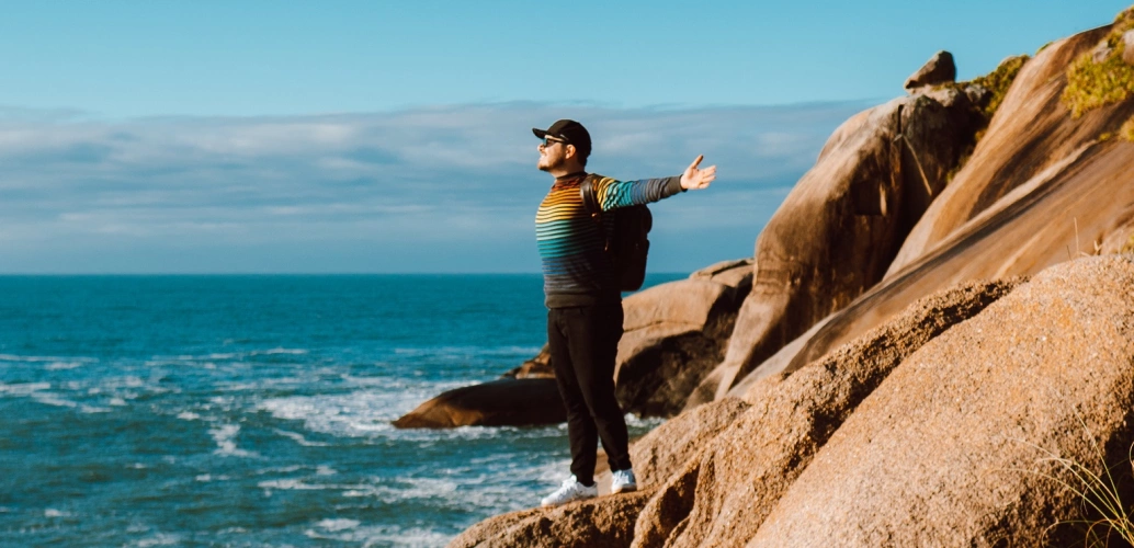 Homem de pé em pedras com braços abertos em direção ao mar