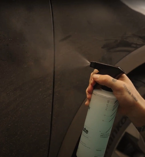 Mão de um homem com tatuagens segurando um borrifador e espirrando o produto na superfície de um carro