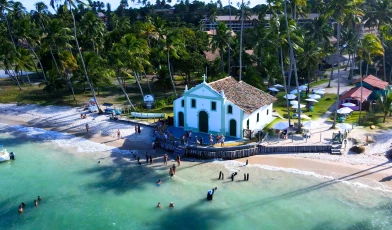 Vista aérea da Praia dos Carneiros. Há uma pequena capela à beira mar, onde estão alguns turistas. Ao fundo, muitos coqueiros e alguns guarda-sóis de praia