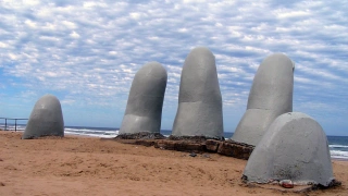 Escultura com as pontas de cinco dedos enormes como se estivessem saindo debaixo da areia da praia