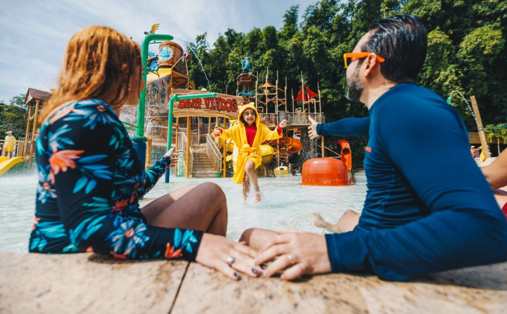 Homem e mulher à frente da foto, criança correndo em direção a eles no centro da imagem. Ao fundo, a estrutura do brinquedo aquático.