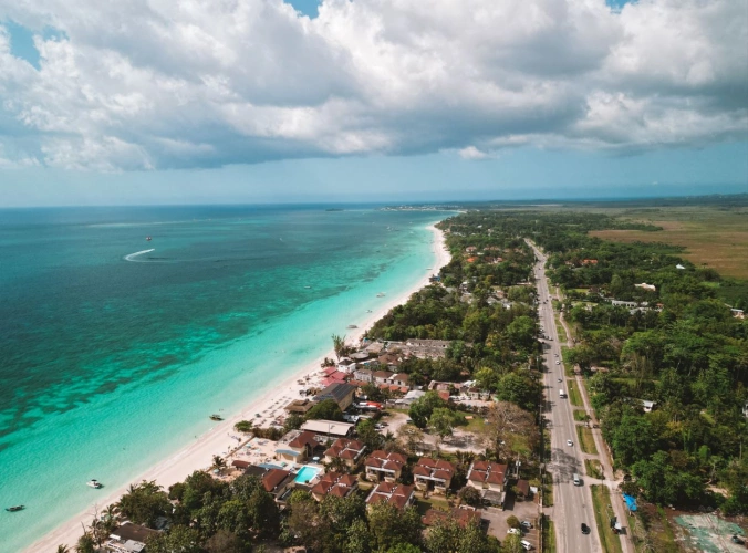 Vista aérea de praia extensa com mar azul-turquesa e areia branca. De frente para a praia, grandes casas e uma estrada em meio à vegetação