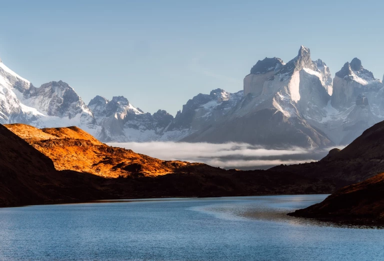 Imagem de lago azul, logo atrás, morros escuros com alguns feixes de luz do sol destacando tons alaranjados. Mais ao fundo uma cadeia de montanhas nevadas e céu nublado em Torres del Paine, na Patagônia chilena.