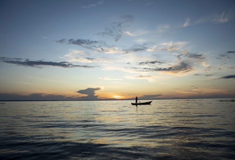 Paisagem de praia deserta com uma pessoa navegando em um barco com a vista do pôr do sol ao fundo da tela.
