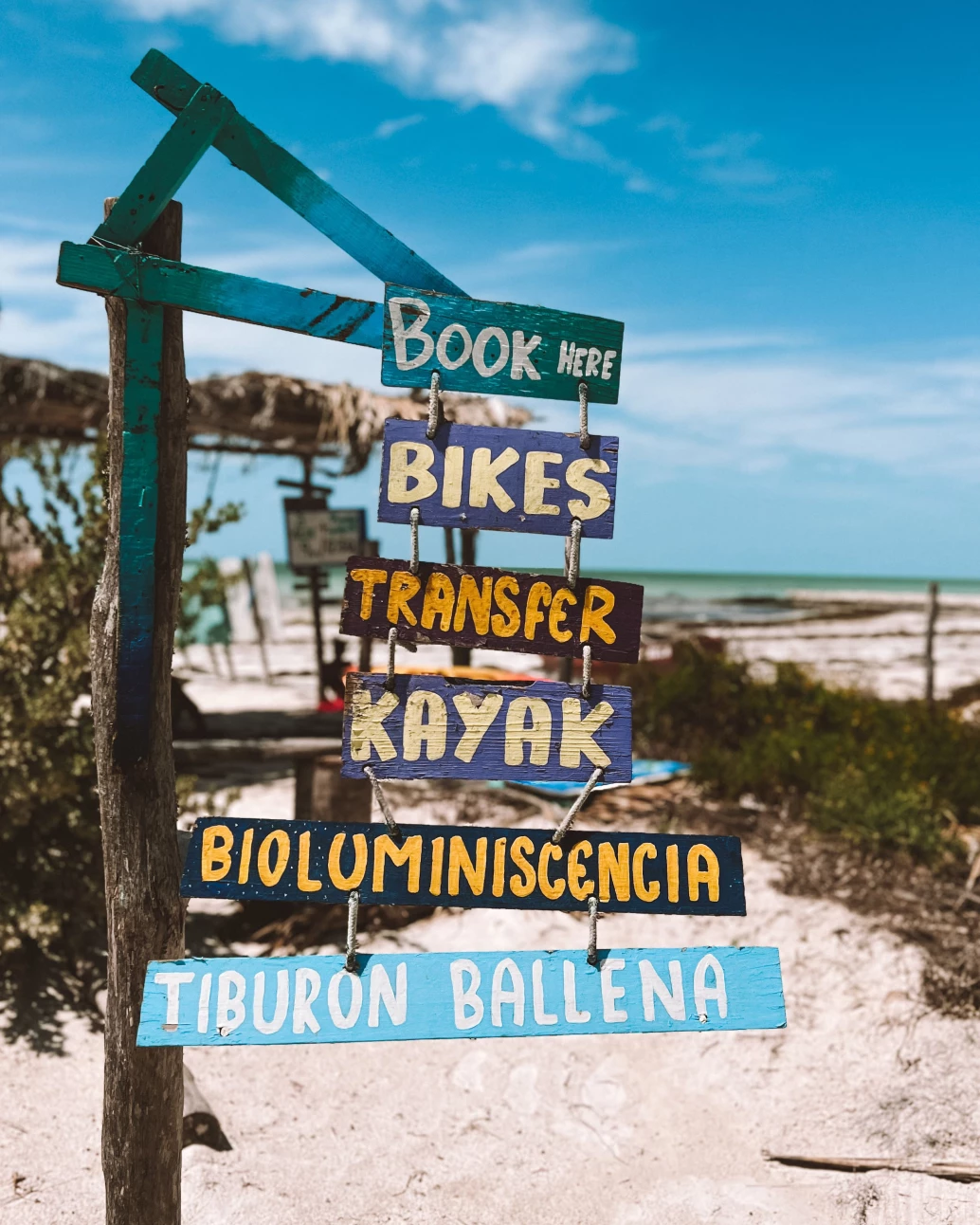 Placa de madeira com diversas atividades escritas, com o mar e a praia ao fundo em Holbox, Quintana Roo.