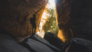 Grandes pedras e paredões de cânion levam a uma abertura na formação rochosa expondo uma fresta de natureza. A silhueta de um homem se destaca ao centro.