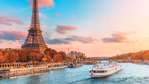 Fotografia da Torre Eiffel ao lado do Rio Sena, por onde passa um barco branco. Céu azul turquesa com um pequeno grupo de pássaros e algumas nuvens.