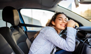 Mulher jovem sorri de olhos fechados e se debruça sobre o volante de um carro.