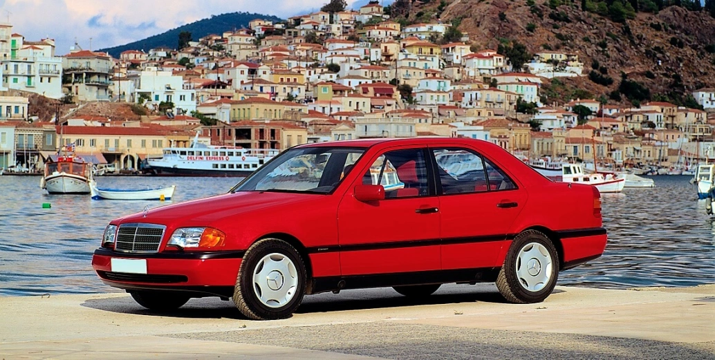 Mercedes-Benz Classe C vermelho estacionado na beira da água, com casas e montanhas ao fundo.