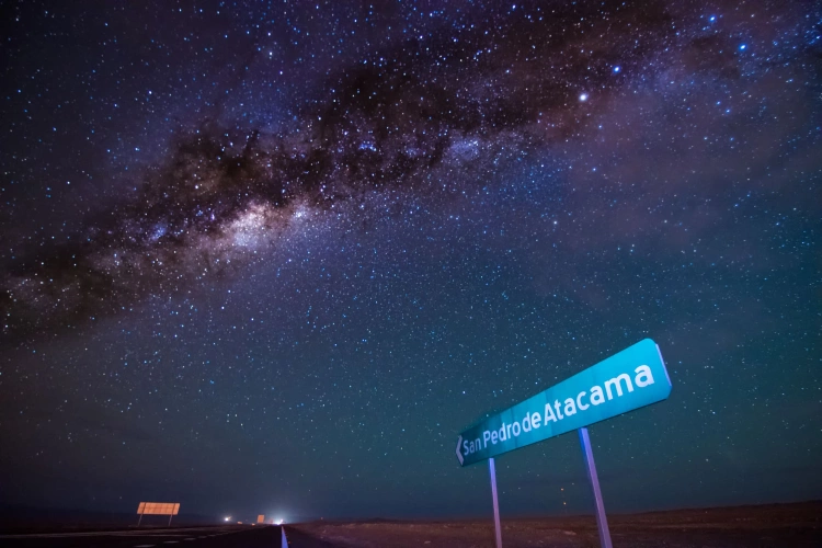 Céu extremamente estrelado, que possibilita ver as constelações e uma galáxia. Uma placa na beira da estrada indica a direção para San Pedro de Atacama