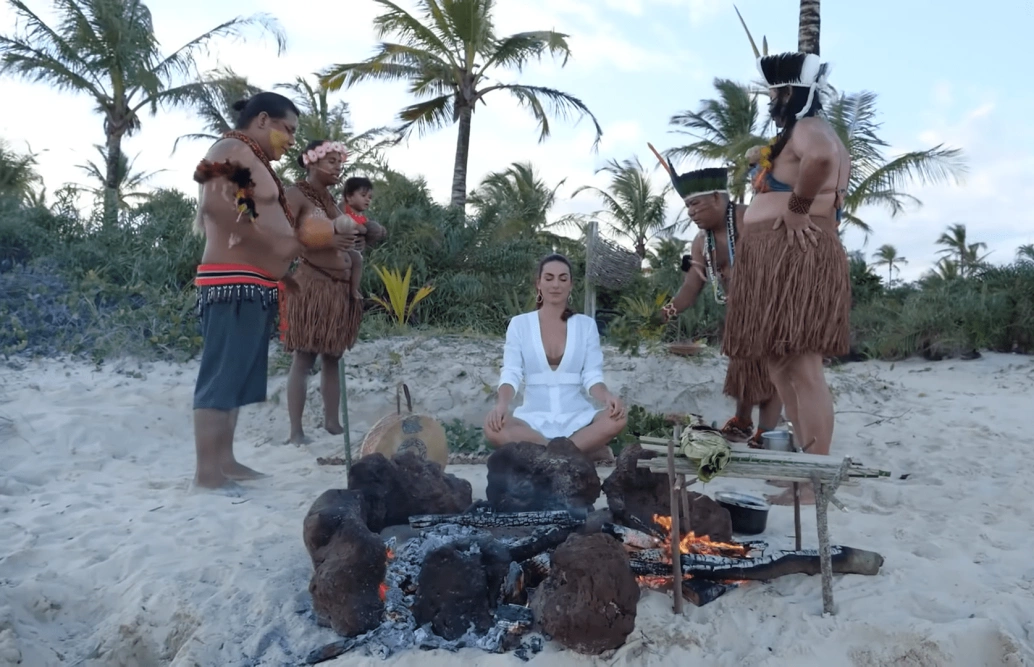 Quatro pessoas de origem indígena ao redor de uma mulher sentada na areia de uma praia