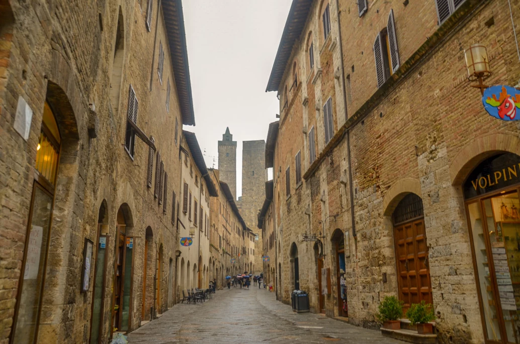 Uma viela em San Gimignano, Toscana, com prédios medievais adornando o caminho. Ao fundo, duas torres se destacam.