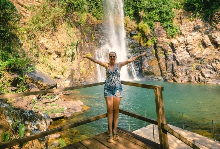 Mulher posa de braços abertos em frente à Cachoeira Serra Azul em dia ensolarado