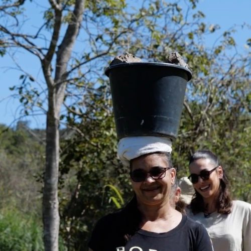 Zezinha Gomes à frente, com balde na cabeça, e  Mel Fronckowiak, ao fundo, posam para foto em meio a vegetação no Vale do Jequitinhonha.