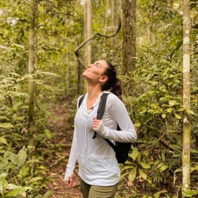 Mulher de mochila nas costas olha para o alto em meio a região florestal