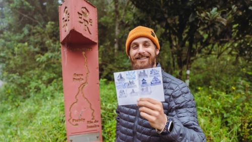 Homem jovem com jaqueta e touca sorrindo para a câmera e segurando um passaporte. Ele está ao lado de um marco da Estrada Real, com vegetação ao fundo