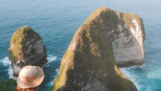 Mulher de costas para a câmera sentada em uma pedra alta observando o mar e uma formação rochosa que lembra a cabeça de um dinossauro