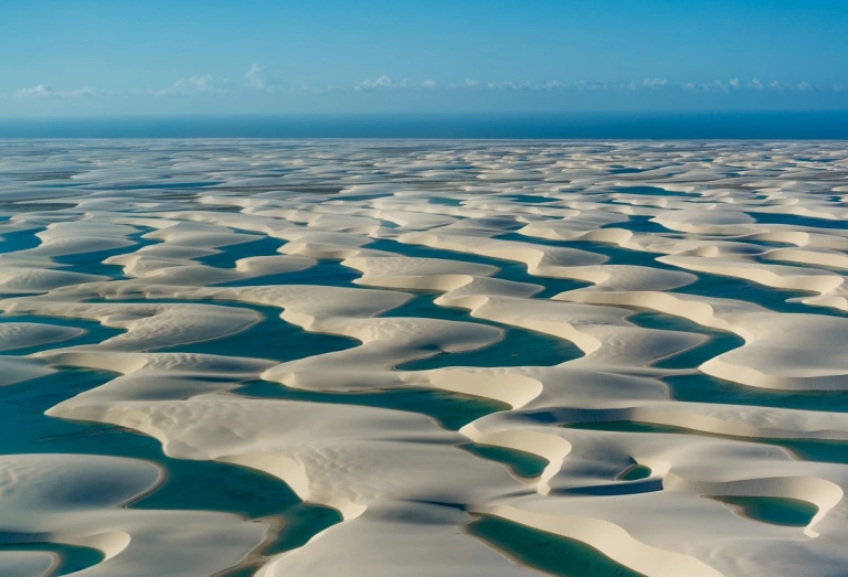 Vista panorâmica do Parque Nacional dos Lençóis Maranhenses com inúmeras lagoas de águas azuis cercadas por dunas de areia em dia ensolarado.