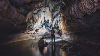 Homem de capacete com lanterna em pé em meio às estruturas de uma caverna escura, iluminando as estalactites