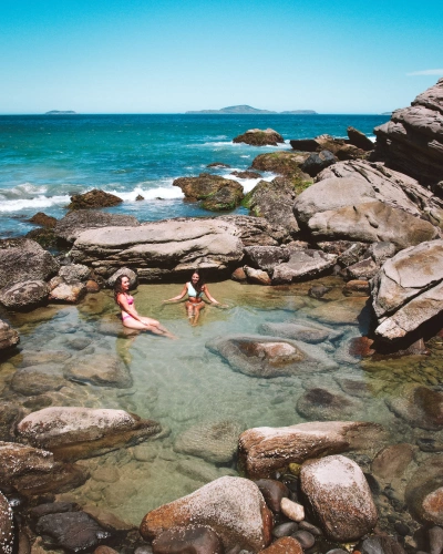 Piscina natural envolta de pedras ao lado do mar. Duas mulheres estão sentadas dentro da água.
