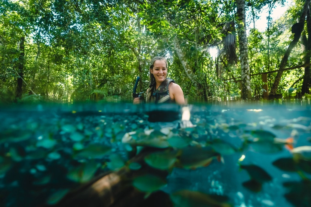 Mulher sorrindo mergulha em lagoa de água azul cristalina com vários peixes submersos
