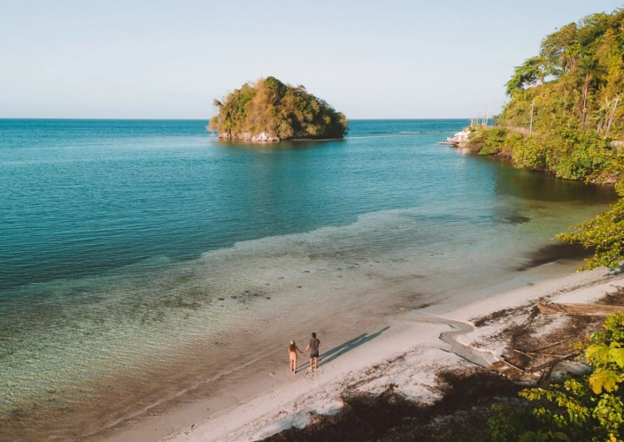 Vista aérea de uma praia e um casal de mãos dadas sobre a areia, avistando uma pequena ilha em meio ao mar