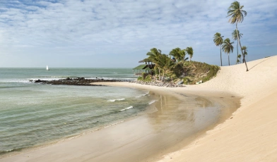 Vista de praia paradisíaca e deserta, mar esverdeado e dunas de areia clara se destacam.