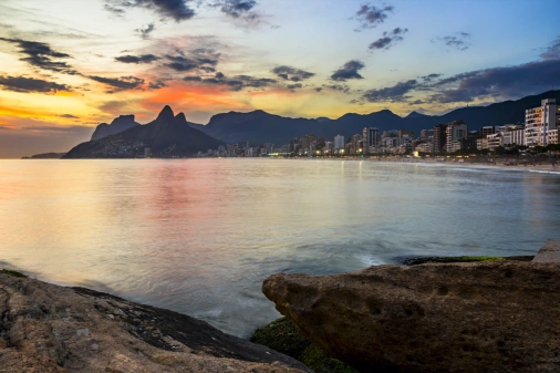 Pôr do sol visto a partir de uma pedra na praia no Rio de Janeiro. O céu alaranjado contrasta com a silhueta das montanhas